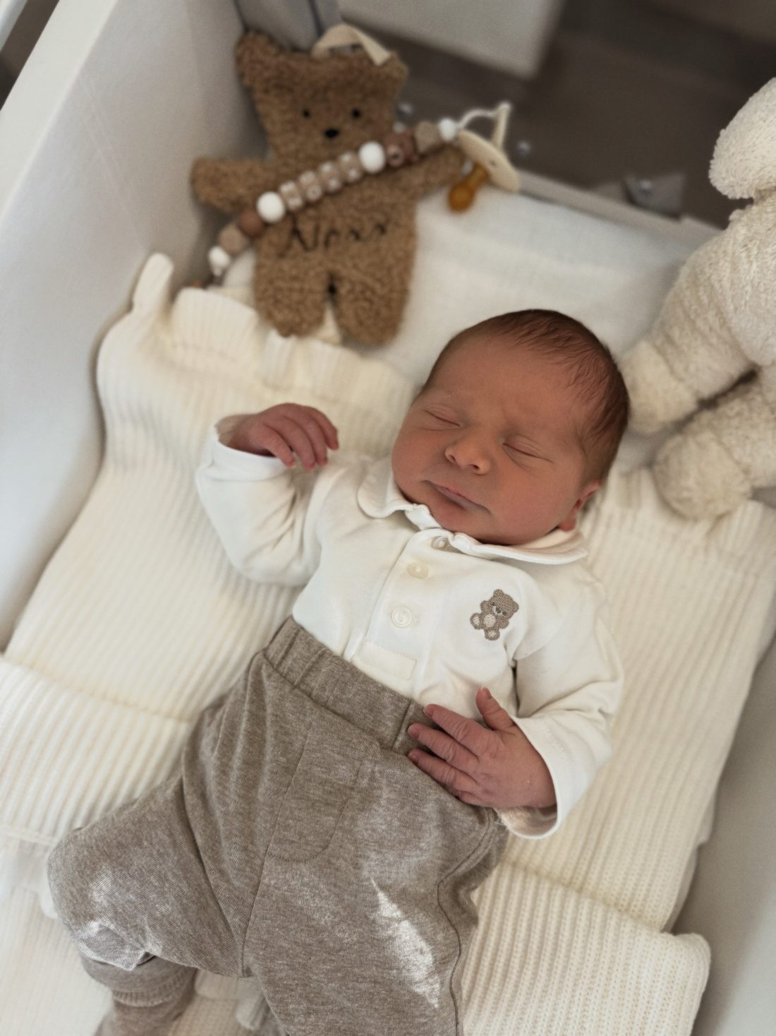 Noxx slaapt in zijn bedje, in een wit bodytje met een beertje en een grijs broekje, met achter hem een knuffelbeer met zijn naam.