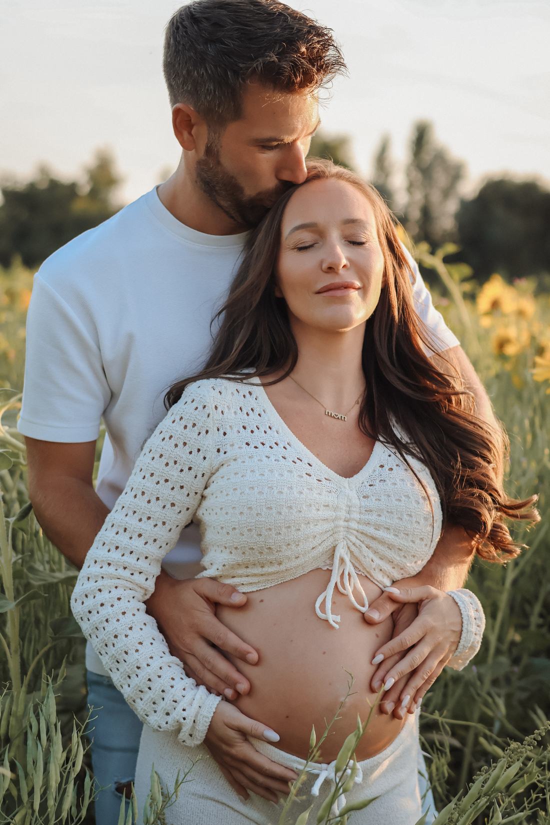 Mama en papa in een bloemenveld bij zonsondergang: papa kust mama op haar hoofd, hun handen samen op haar buik.